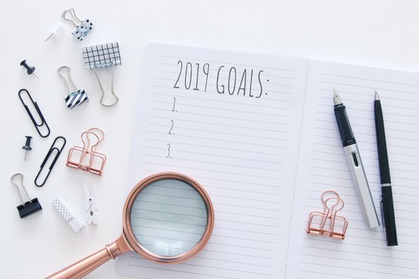 Top view 2019 goals list with notebook over white wooden desk.